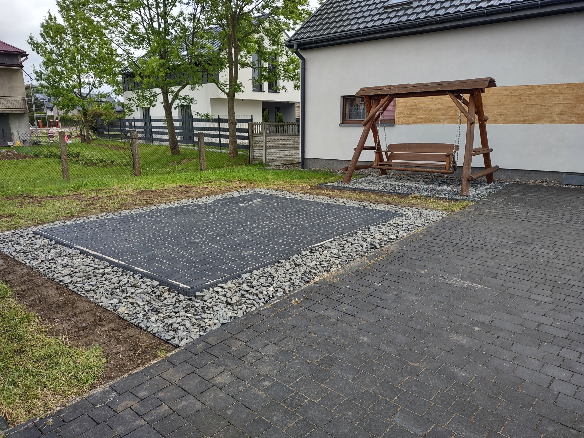 Wykończona nawierzchnia z ciemnej kostki brukowej, otoczona szarym żwirem, z drewnianą huśtawką w tle, na tle białego domu z szarą dachówką. Widok z góry.