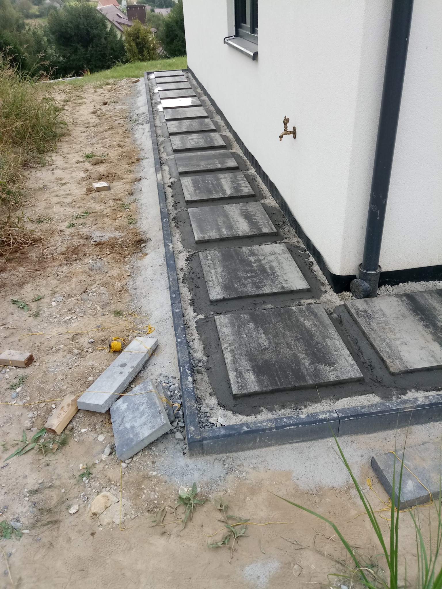 Ścieżka z płyt betonowych w trakcie montażu przy białej ścianie budynku z kranem. Widoczne narzędzia i materiały budowlane.