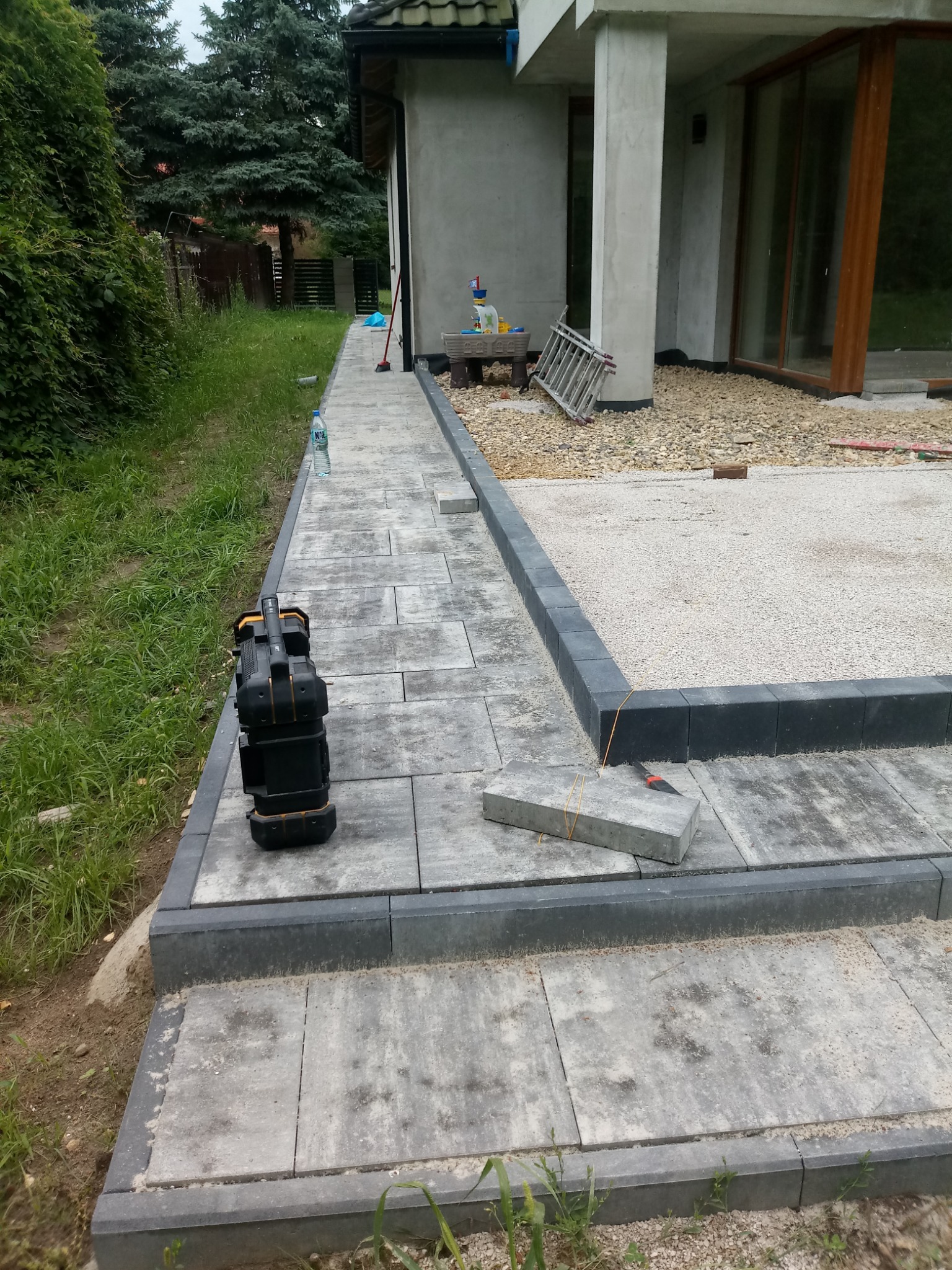 Ułożona ścieżka z szarej kostki brukowej prowadząca wzdłuż nowoczesnego domu z dużymi oknami. Widoczne narzędzia i materiały budowlane. Etap prac wykończeniowych.