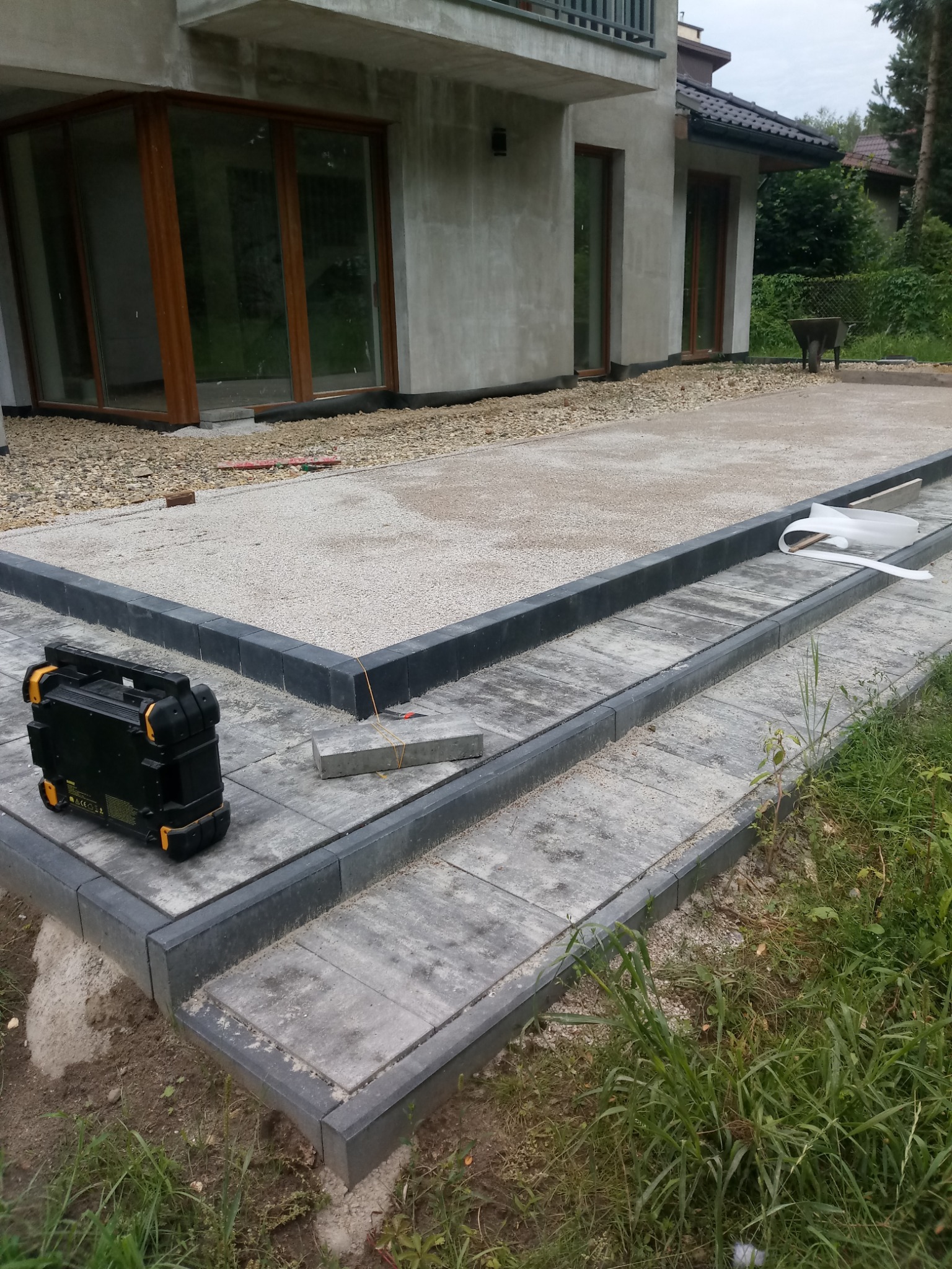 Ułożona szara kostka brukowa z ciemnymi obrzeżami na podwórku domu. Widoczny sprzęt budowlany i elementy przygotowane do dalszych prac, częściowo podsypane kamieniami.