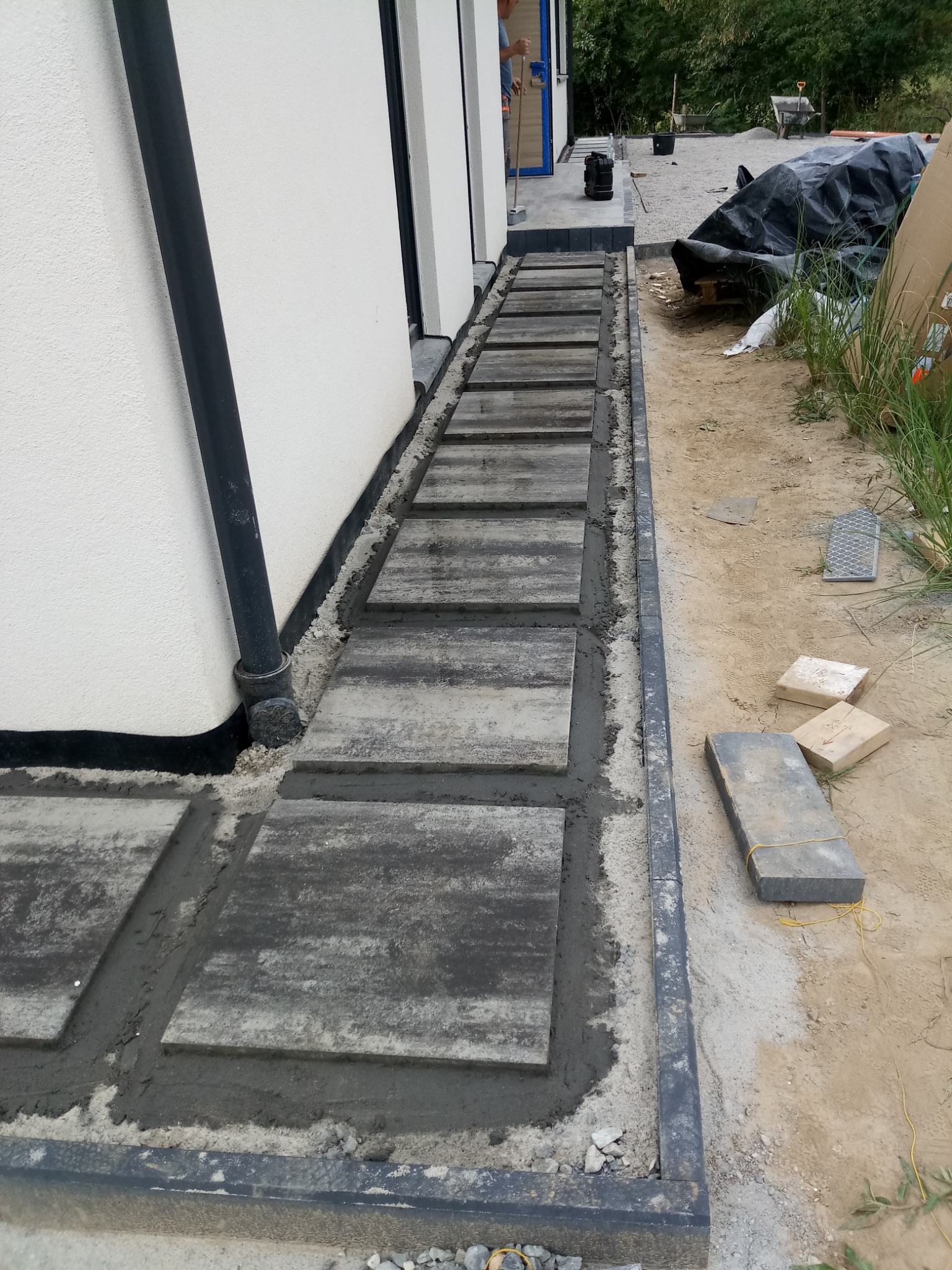 Ścieżka z szarych płyt betonowych układana przy białej ścianie budynku. Widoczne etapy prac, świeży cement między płytami i obrzeżem. W tle narzędzia i teren budowy.
