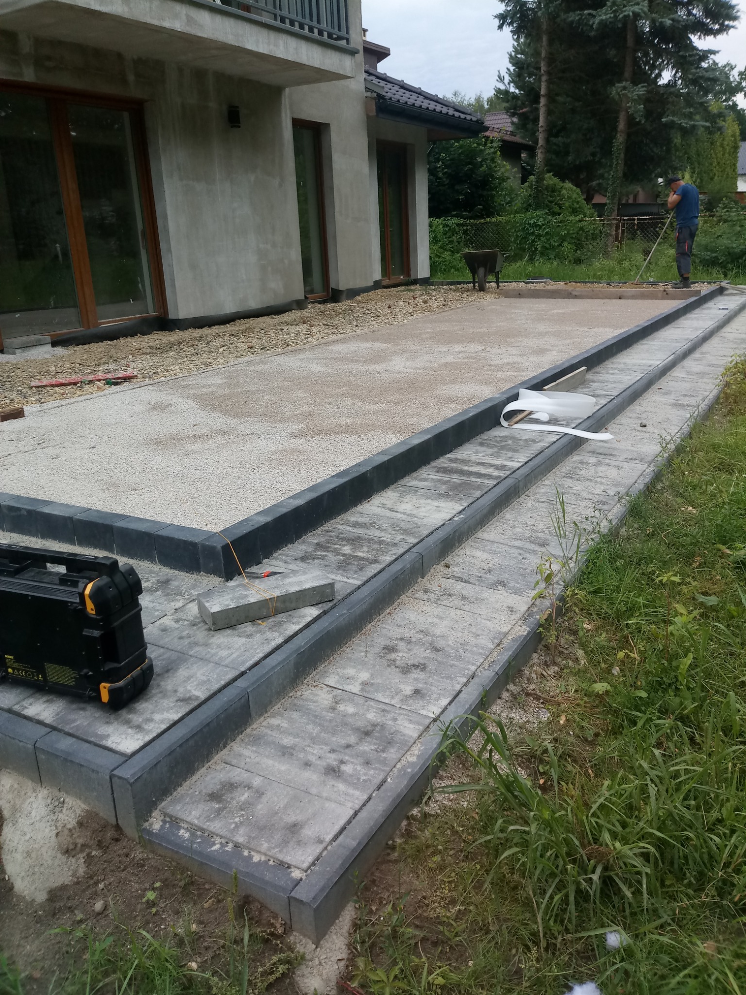 Etap prac: przygotowanie podłoża i obrzeży z kostki granitowej, widoczny piasek, narzędzia i bryła budynku w tle. Prace brukarskie w toku.