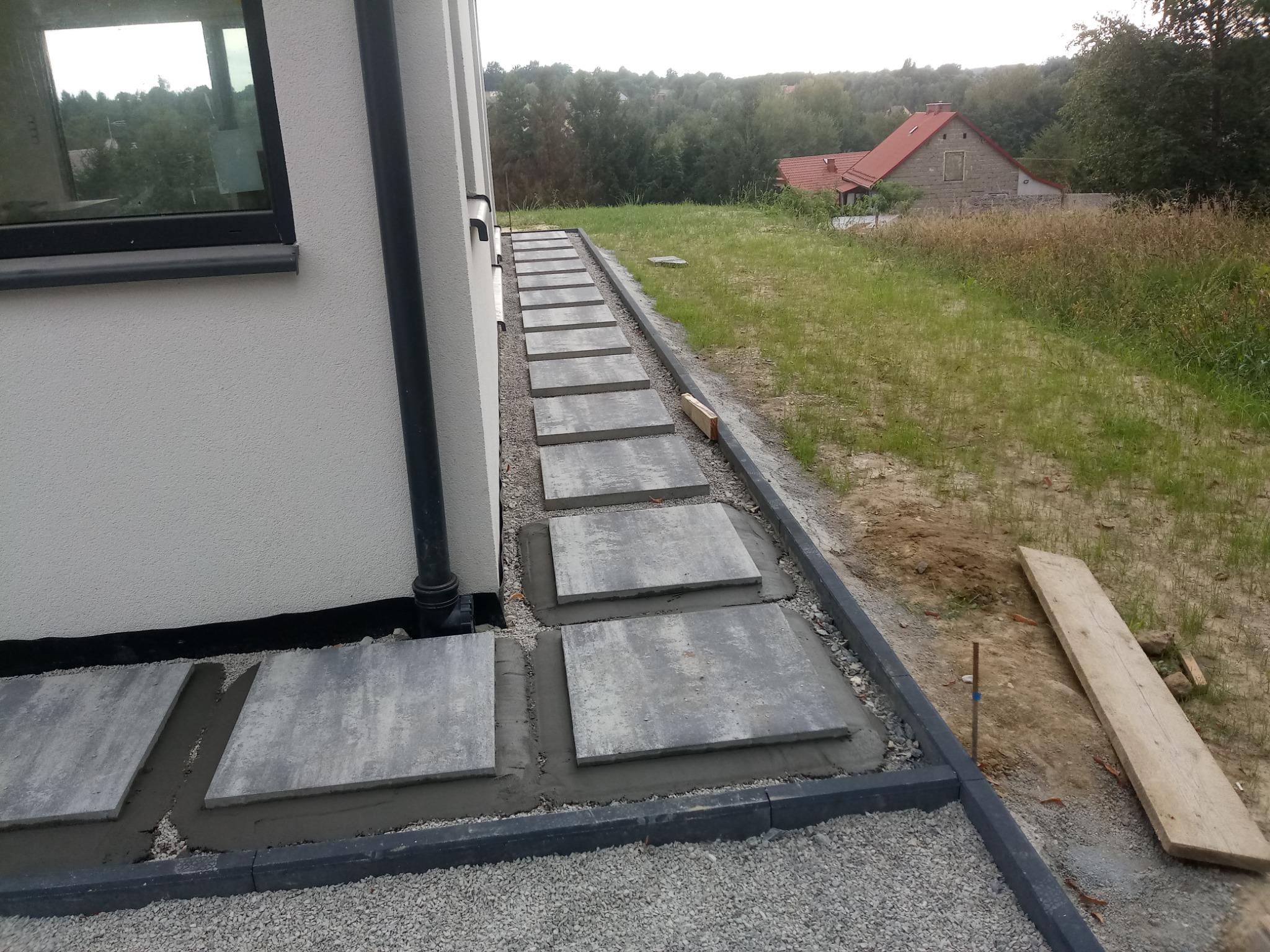 Ścieżka z kwadratowych płyt betonowych ułożona wzdłuż budynku w Siedlcu, obrzeża wykończone kostką. Widoczny trawnik i dom w tle.