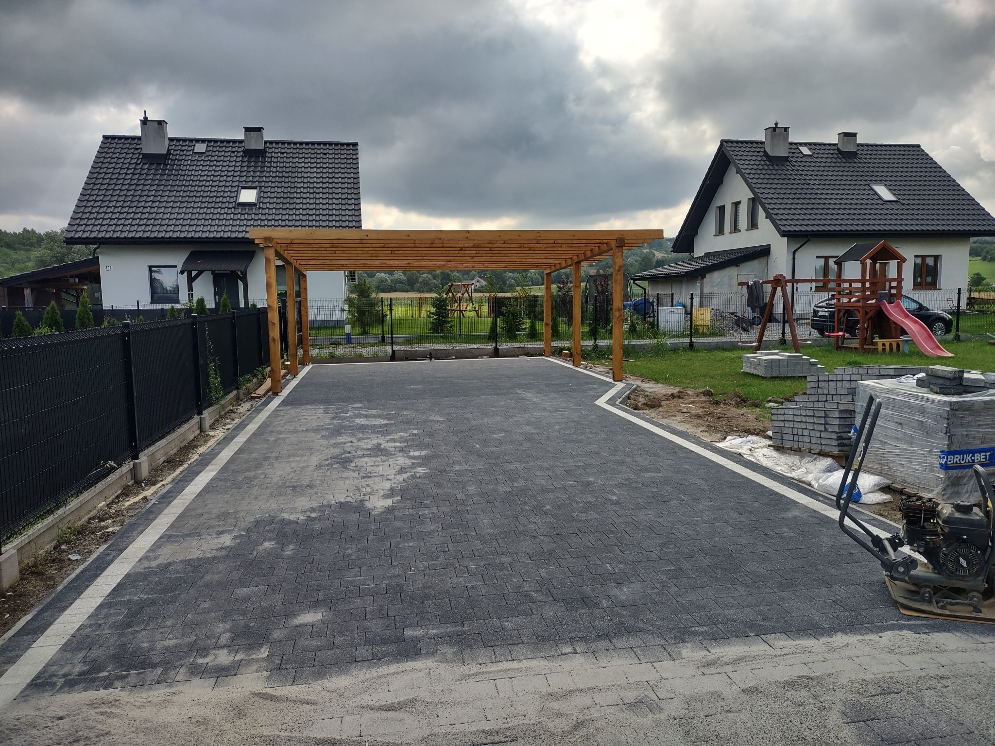 Ułożona, grafitowa kostka brukowa tworzy podjazd pod drewnianą wiatą, otoczony zielenią i domami. Widoczna ubijarka i paleta z kostką.