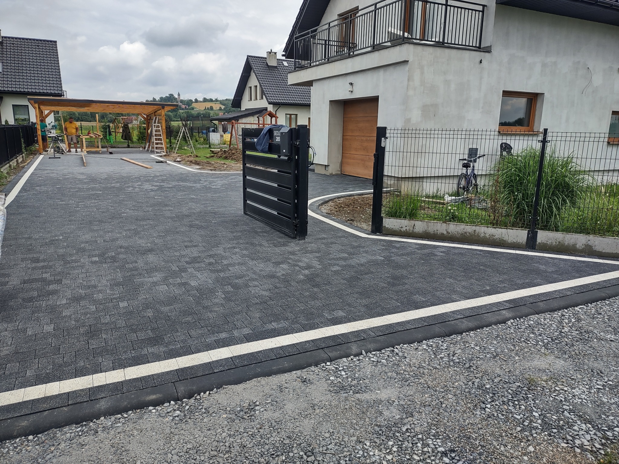 Ukończony podjazd z szarej kostki brukowej z białymi pasami, czarna brama wjazdowa, widoczny dom z garażem i częściowo zadaszenie w trakcie budowy.