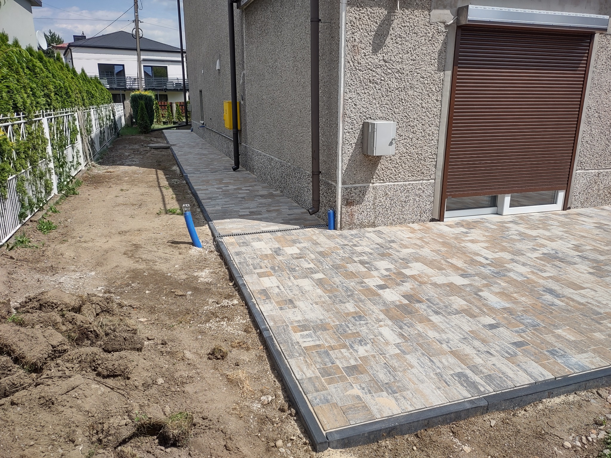 Ułożona kostka brukowa w odcieniach szarości i beżu, obrzeża z ciemnej kostki. Częściowo wykończony teren przy domu z roletami zewnętrznymi.