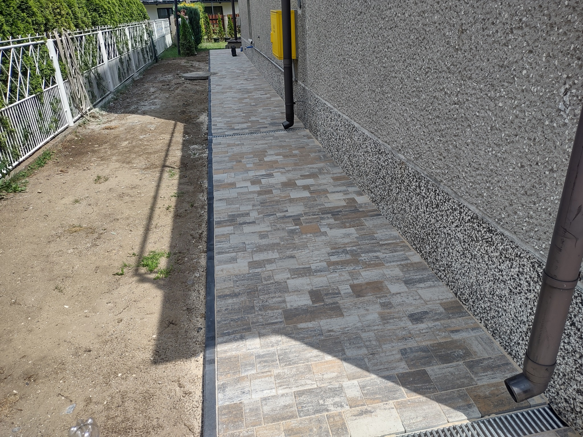 Ułożona kostka brukowa wzdłuż ściany budynku, z widocznym odwodnieniem liniowym i fragmentem trawnika. Kostka w odcieniach szarości i beżu.