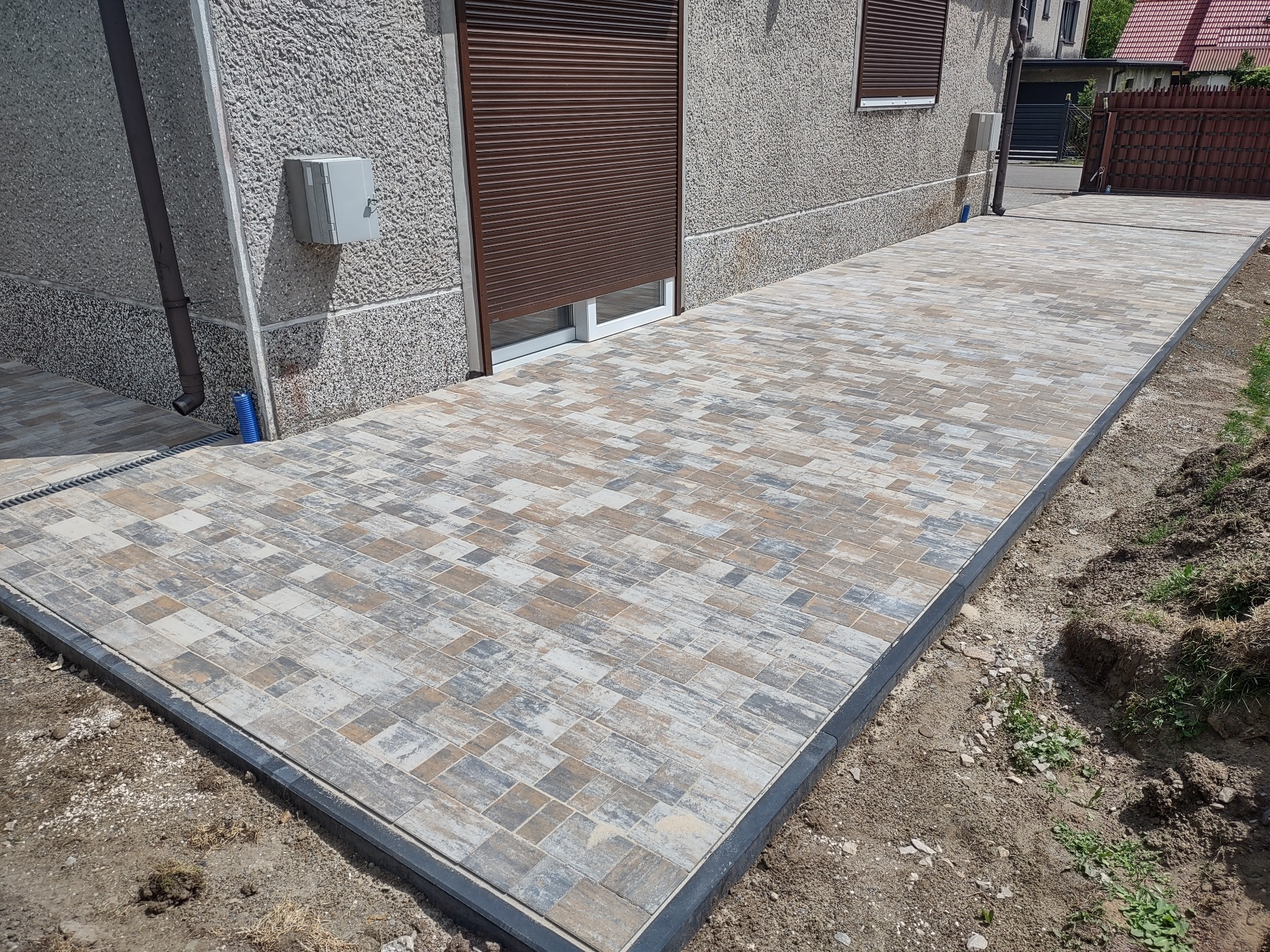 Ścieżka z kostki brukowej o nieregularnym wzorze, obramowana czarnym obrzeżem, prowadząca wzdłuż elewacji domu z brązowymi roletami okiennymi.