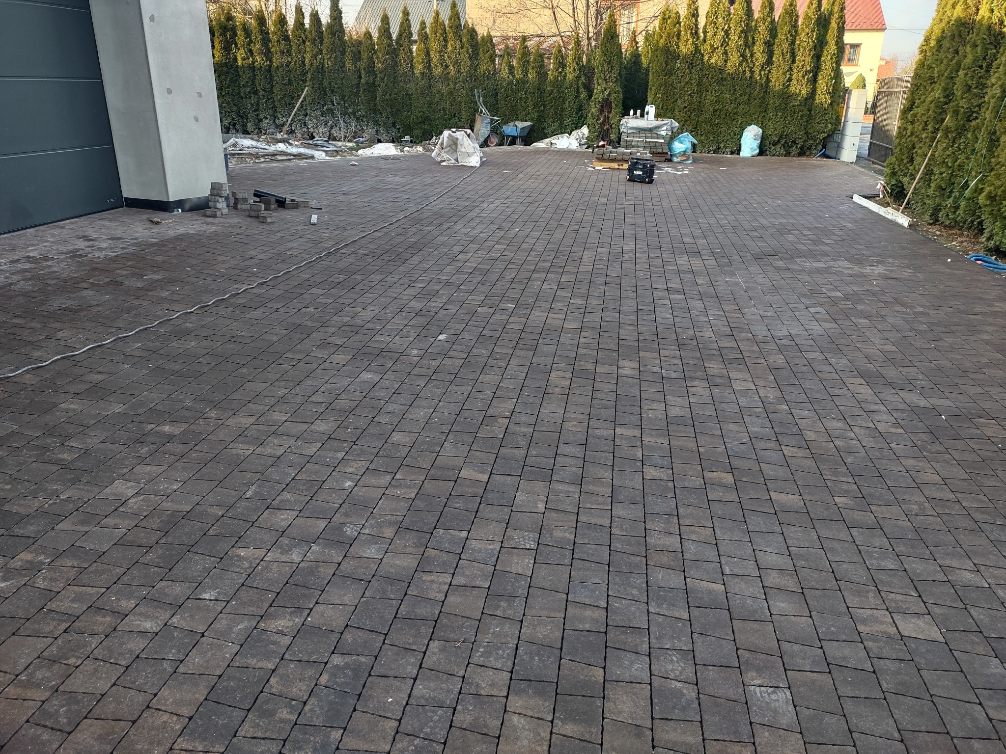 Ułożona, brązowa kostka brukowa na podjeździe, z widocznymi narzędziami i materiałami budowlanymi w tle, otoczona tujami. Widok z perspektywy.
