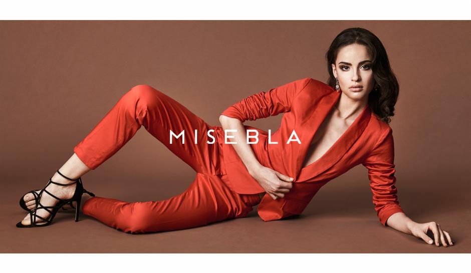 Modelka w czerwonym garniturze i czarnych szpilkach, pozująca na brązowym tle w Jarocinie, prezentuje odważną stylizację.