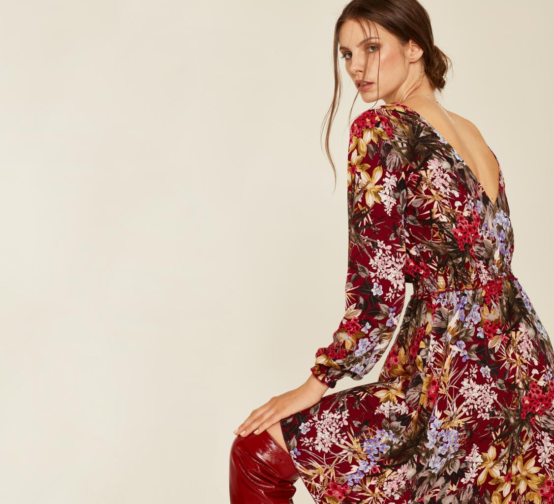 Kobieta spogląda przez ramię, ubrana w kwiecistą sukienkę z długim rękawem i czerwone lakierowane kozaki za kolano.