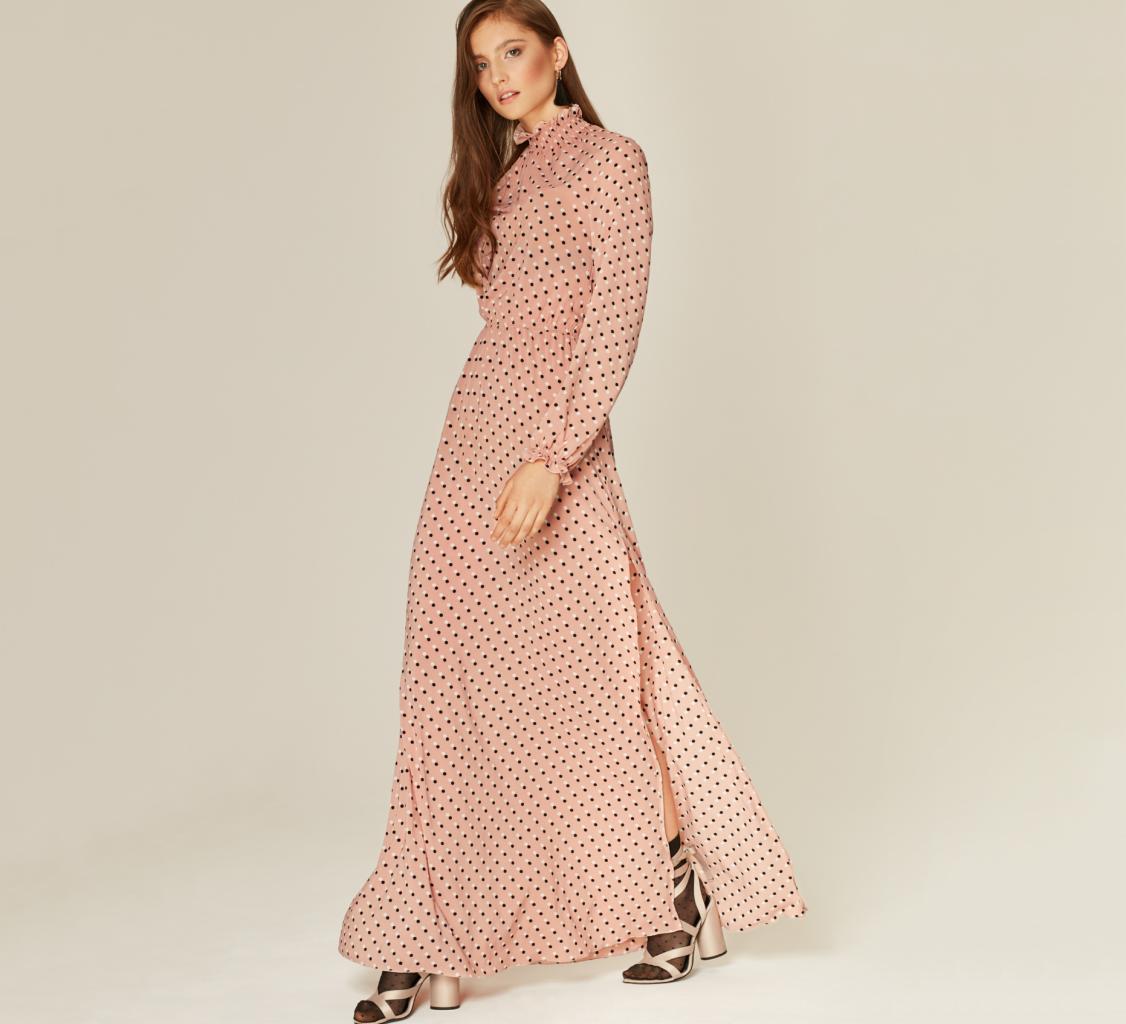 Modelka w długiej, pudrowo różowej sukience w czarne kropki z rozcięciem na nodze i stójką, pozuje w złotych sandałach na obcasie.