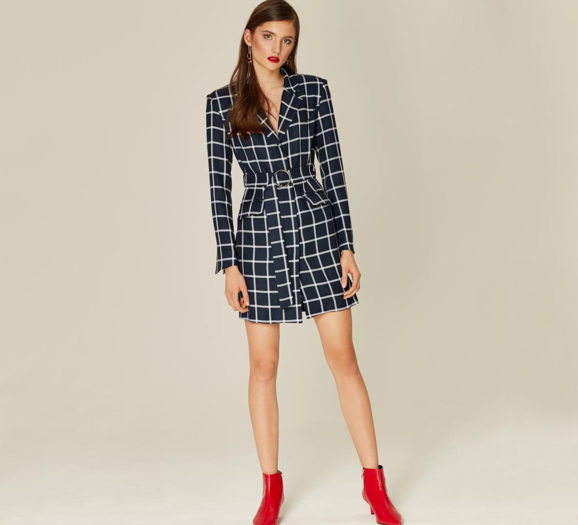 Modelka prezentuje granatową sukienkę w białą kratę z paskiem, uzupełnioną czerwonymi botkami na obcasie i długimi kolczykami.