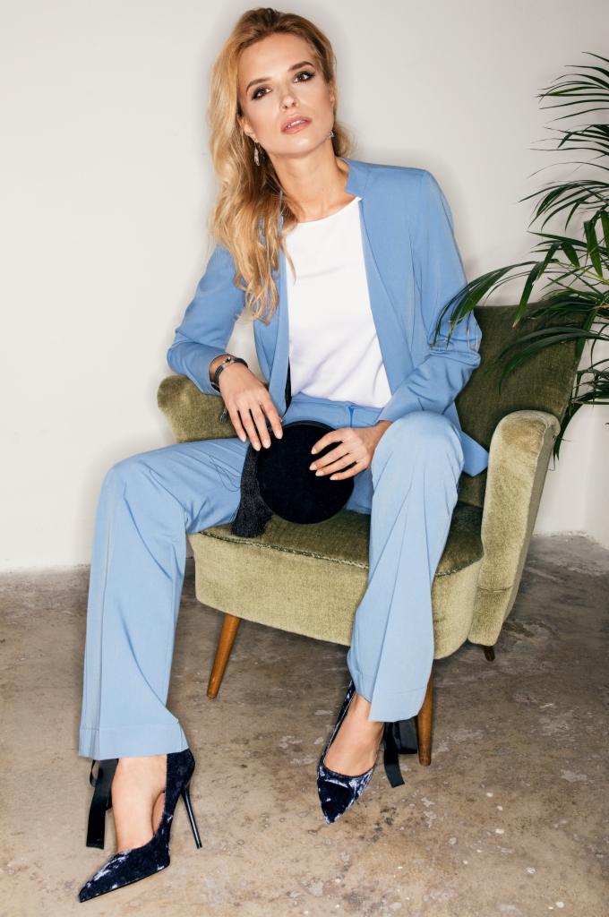 Kobieta w błękitnym garniturze i aksamitnych szpilkach siedzi w fotelu, trzymając małą okrągłą torebkę z frędzlami.