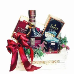 Elegancki kosz prezentowy z butelką alkoholu, czekoladkami i herbatą, ozdobiony czerwoną kokardą, umieszczony w białym, drewnianym koszyku.