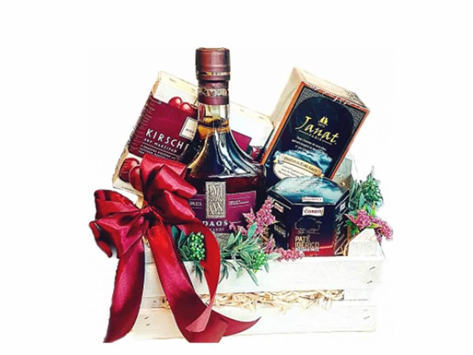 Elegancki kosz prezentowy z butelką alkoholu, czekoladkami i herbatą, ozdobiony czerwoną kokardą, umieszczony w białym, drewnianym koszyku.
