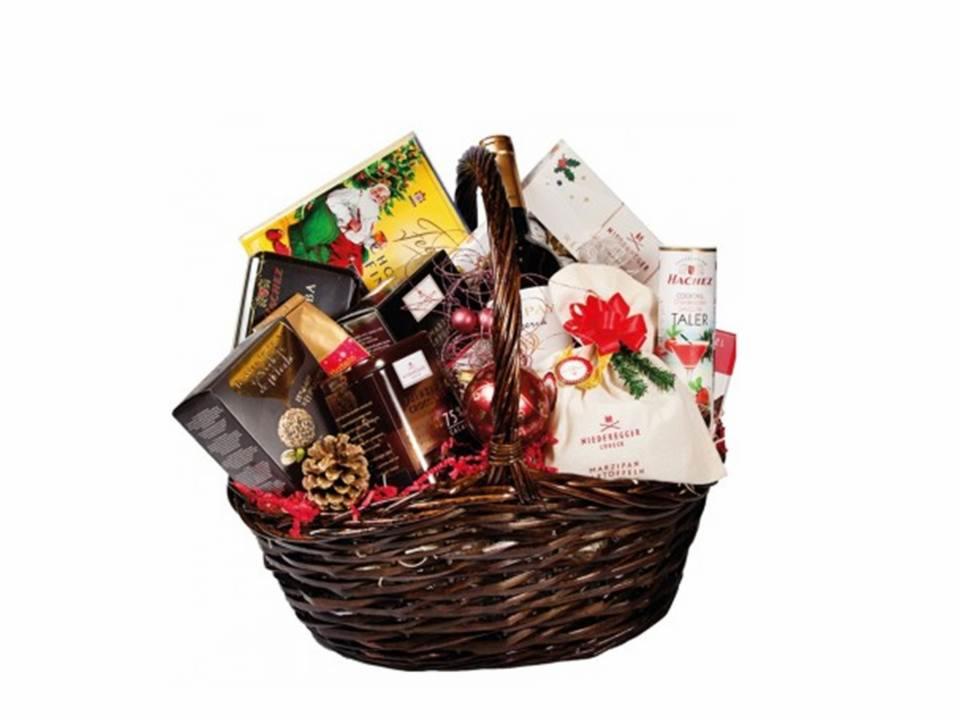 Kosz prezentowy z różnorodnymi słodyczami, czekoladami i napojami, udekorowany czerwoną wstążką i szyszką, na białym tle.