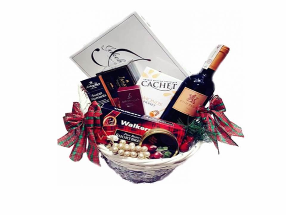 Elegancki kosz prezentowy z butelką czerwonego wina Fortius, czekoladkami Cachet, Walker Shortbread i innymi słodyczami, ozdobiony czerwoną kokardą w kratę i gałązkami świerku.