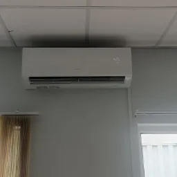 Biały klimatyzator zamontowany na szarej ścianie w pomieszczeniu biurowym, widoczne okna z żaluzjami i fragment sufitu kasetonowego.