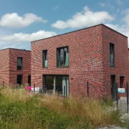 Dwa nowoczesne, prostopadłościenne domy z czerwonej cegły z dużymi oknami, widoczne z poziomu gruntu, z wysoką trawą na pierwszym planie i błękitnym niebem z chmurami w tle.