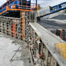 Betonowanie fundamentów pod mury oporowe przy użyciu szalunków systemowych, w tle widoczny sprzęt do transportu kruszywa.