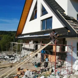 Pracownik na rusztowaniu montuje obróbkę blacharską dachu domu jednorodzinnego z widoczną konstrukcją więźby i elementami ocieplenia, na pierwszym planie gruz i materiały budowlane.