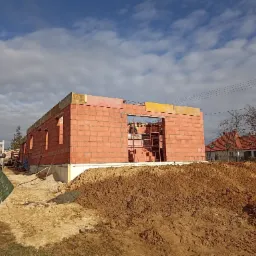 Wznoszenie ścian parterowego domu z czerwonej cegły ceramicznej, widoczne rusztowanie w otworze okiennym i sterta ziemi na pierwszym planie, w tle dom z czerwoną dachówką.