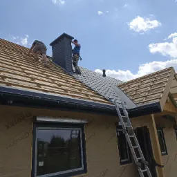 Montaż czarnego komina systemowego na dachu z szarej blachodachówki, widoczna więźba dachowa i dwaj dekarze pracujący na dachu, drabina oparta o budynek w trakcie budowy.