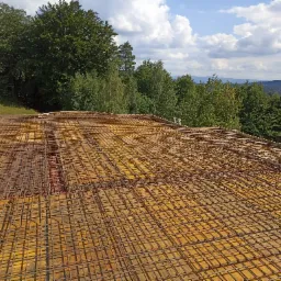 Zbrojenie płyty fundamentowej na żółtym szalunku, widok z góry na tle zieleni drzew i gór w oddali pod błękitnym niebem.