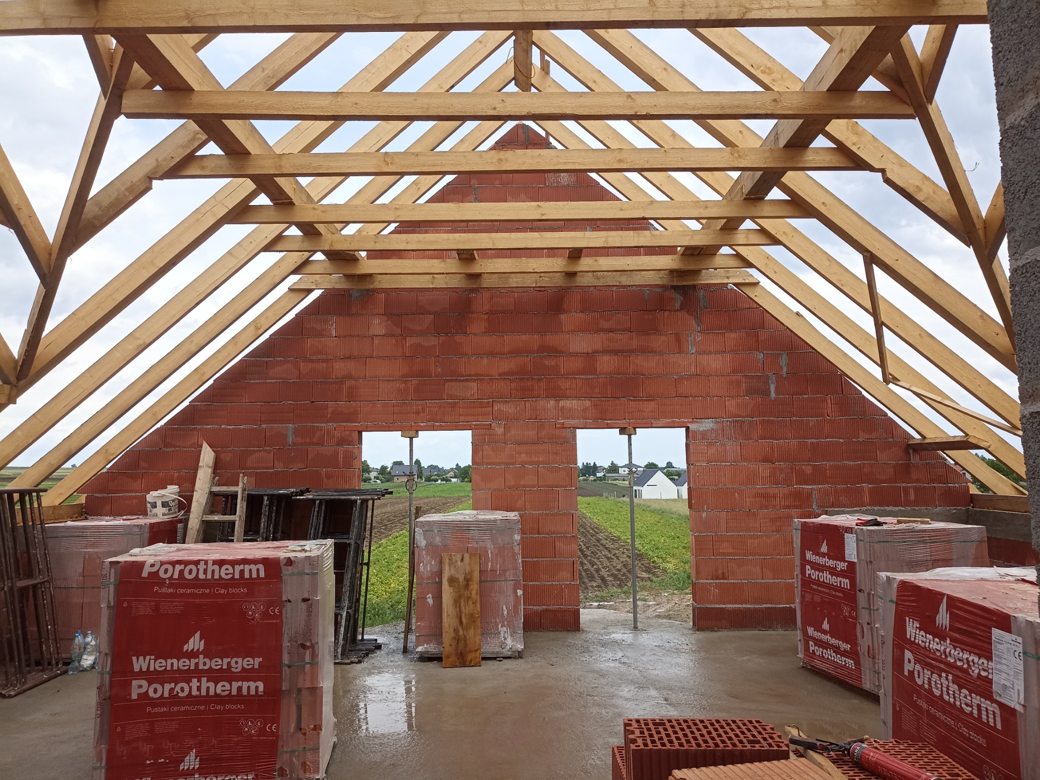 Surowy stan domu murowanego z widoczną więźbą dachową i paletami pustaków Porotherm, widok przez otwory okienne na pola i zabudowania.