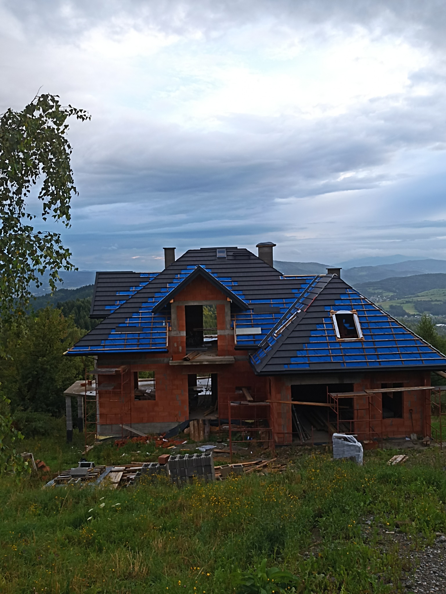 Budowa domu z cegły, z częściowo położonym dachem z czarnej dachówki, zabezpieczonym niebieską folią, na wzgórzu z widokiem na zalesione góry w pochmurny dzień.