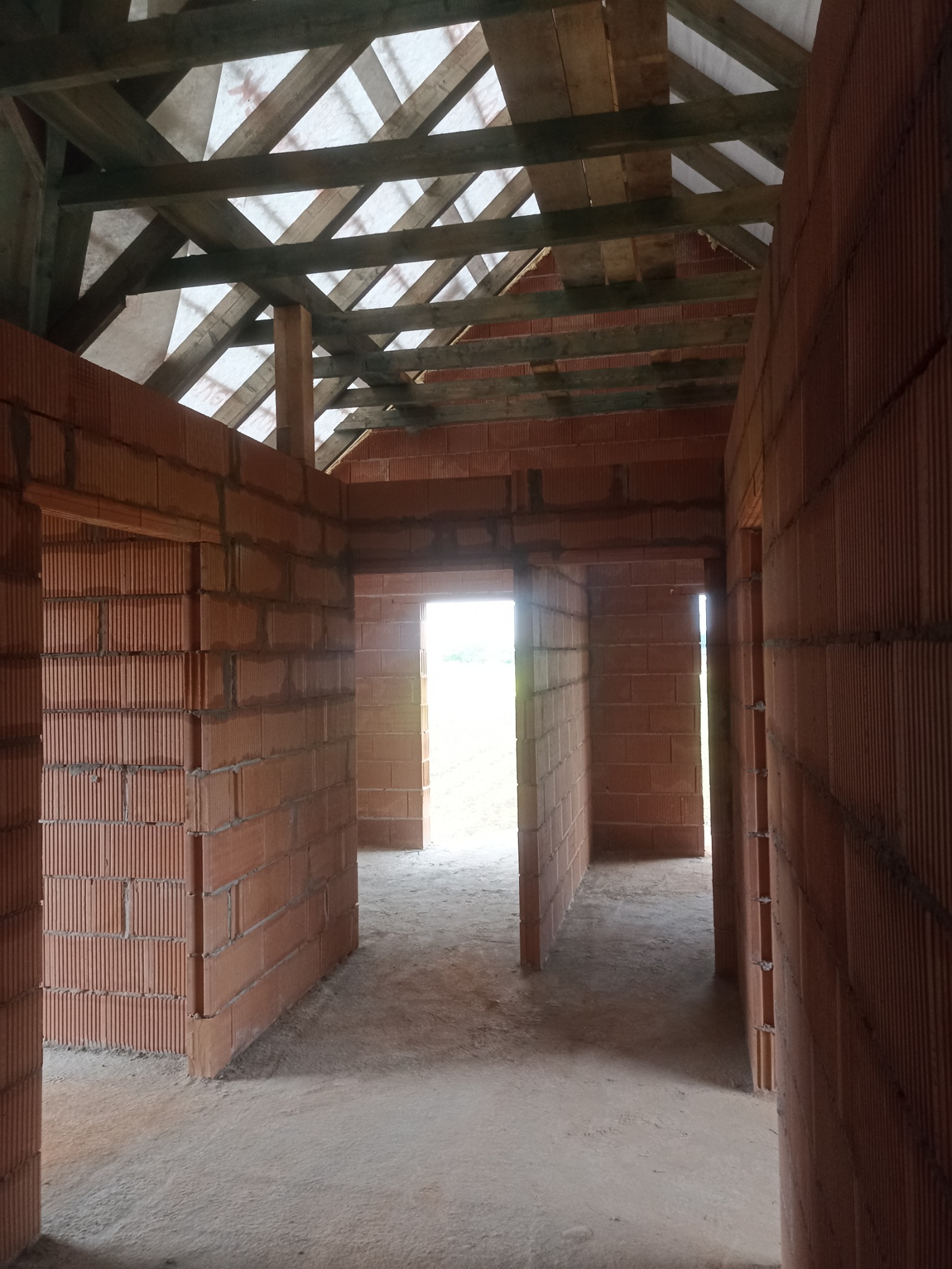 Wnętrze surowego domu w trakcie budowy, widoczne ściany z czerwonej cegły, surowa podłoga i drewniana konstrukcja dachu z widoczną membraną dachową.