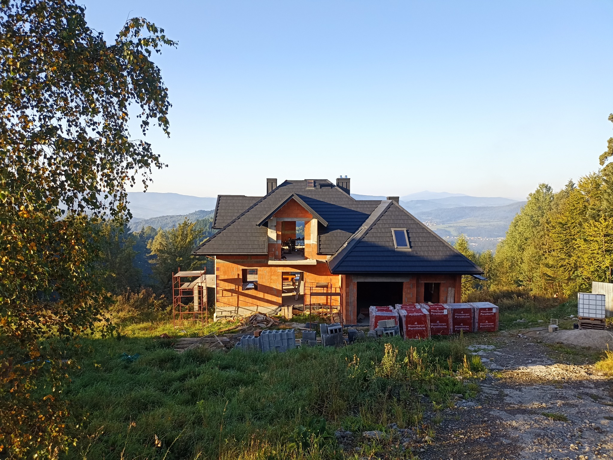 Surowy stan domu murowanego z częściowo ukończonym dachem z dachówki ceramicznej, widoczne materiały budowlane i rusztowania, malowniczy krajobraz górski w tle.