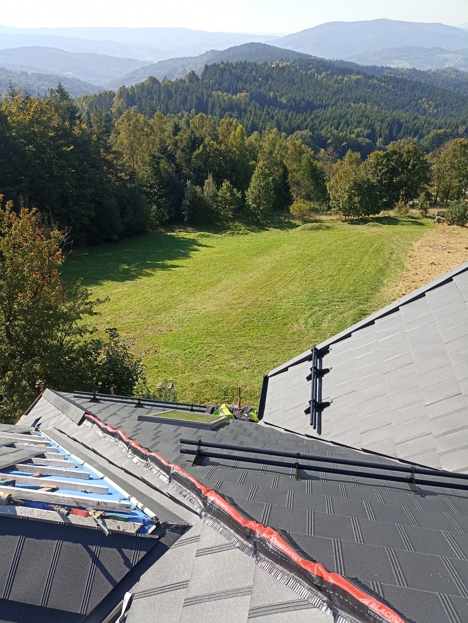 Montaż blachodachówki na dachu z widokiem na zalesione wzgórza w oddali, widoczne narzędzia i materiały budowlane, częściowo ukończona konstrukcja.