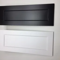 Dwie próbki frontów meblowych: czarny matowy i biały z frezowanym obramowaniem, prezentowane na jasnym tle ściany.