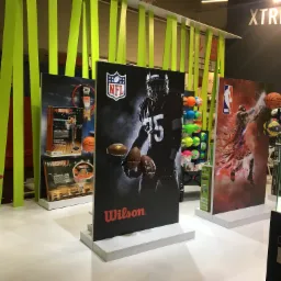 Ekspozycja w sklepie sportowym z planszami reklamowymi przedstawiającymi sportowców i sprzęt sportowy, w tle neonowe zielone dekoracje i automat do popcornu.