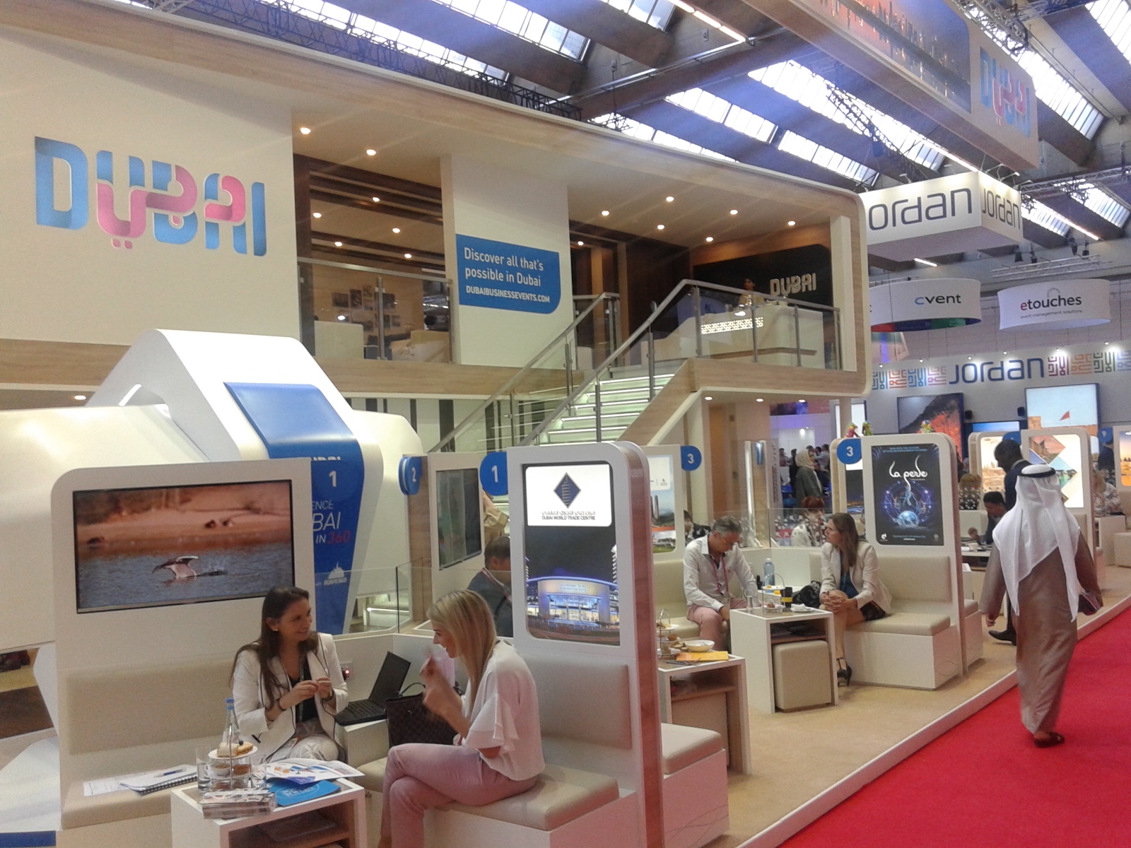 Stoisko promocyjne Dubaju na targach, z osobami siedzącymi na białych kanapach i oglądającymi prezentacje wideo.