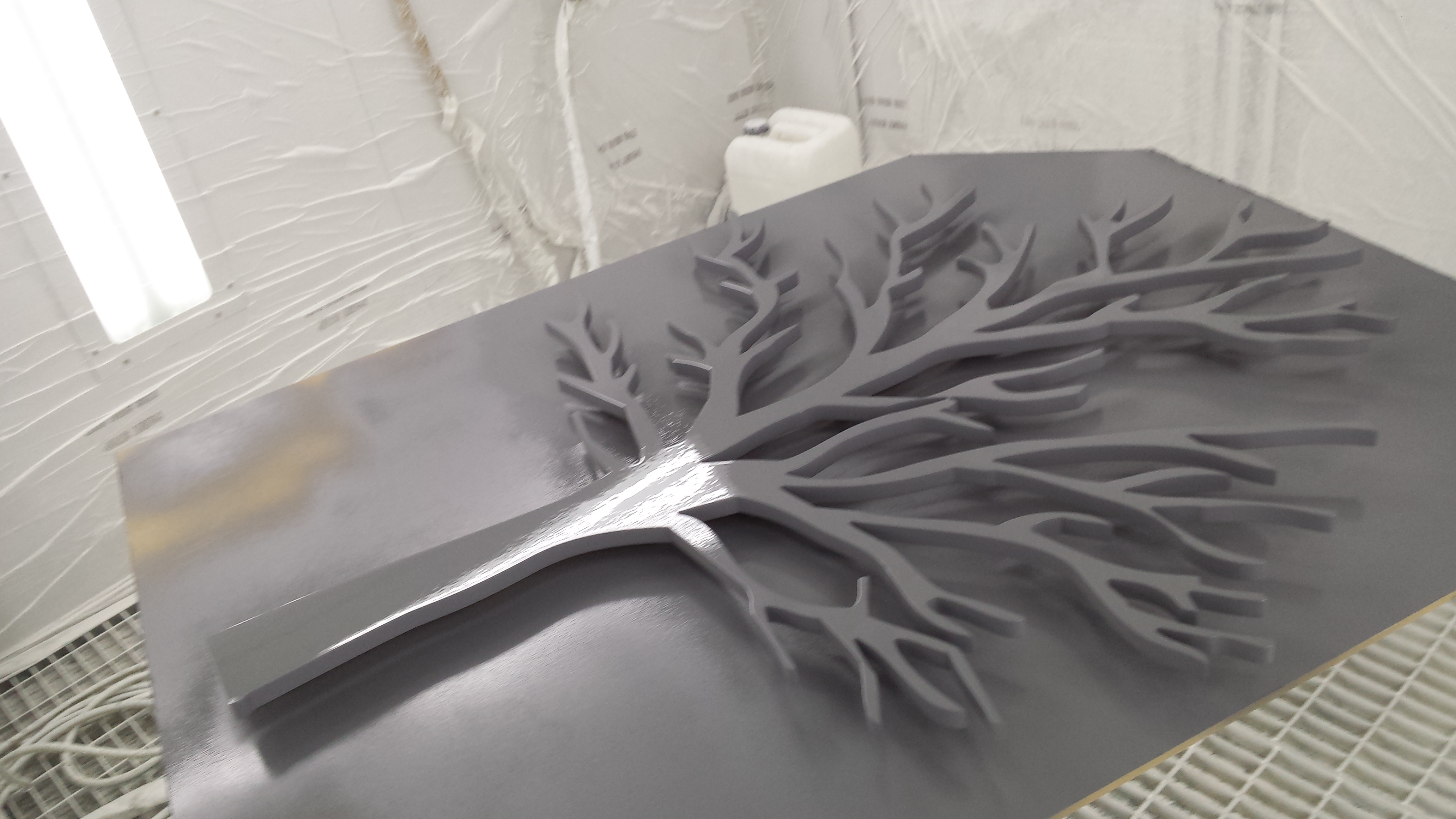 Szary, drewniany element dekoracyjny w kształcie drzewa, leżący na lakierowanej powierzchni w pomieszczeniu warsztatowym.