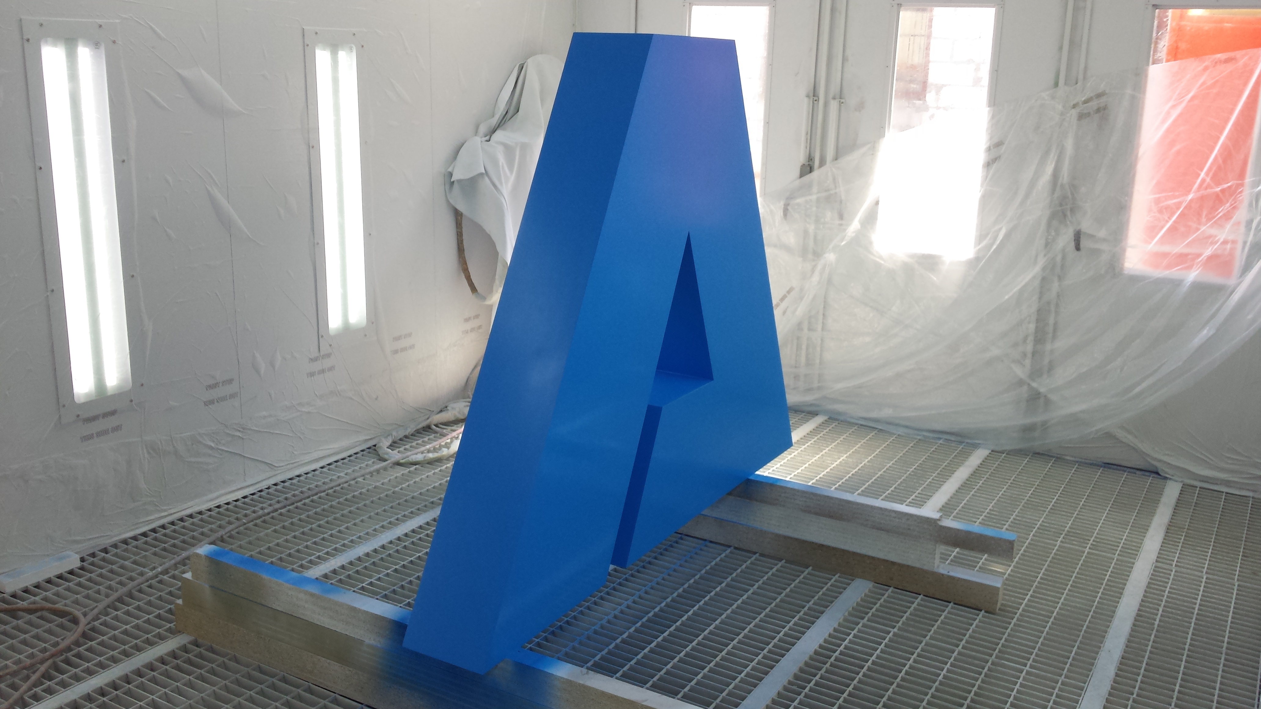 Niebieska, przestrzenna litera A, świeżo pomalowana, ustawiona w komorze lakierniczej na metalowej kratownicy. W tle widoczne okna i folia ochronna.