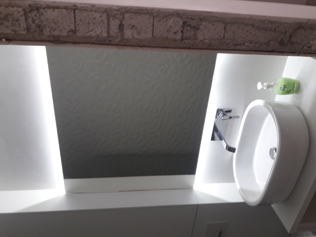 Biała, owalna umywalka nablatowa z chromowaną baterią, podświetlona od góry listwą LED, z dozownikiem mydła w tle, na tle ściany z subtelnym wzorem.