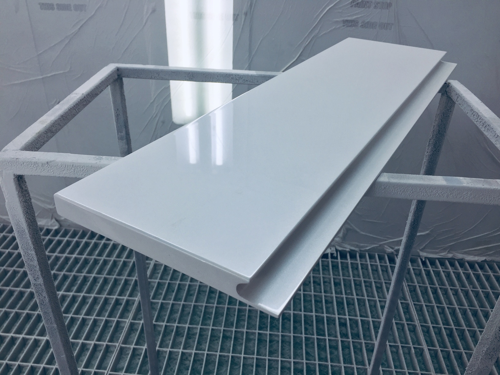 Biały, lakierowany blat na metalowym stelażu w lakierni, widoczne refleksy światła na powierzchni, kratka ściekowa w tle.