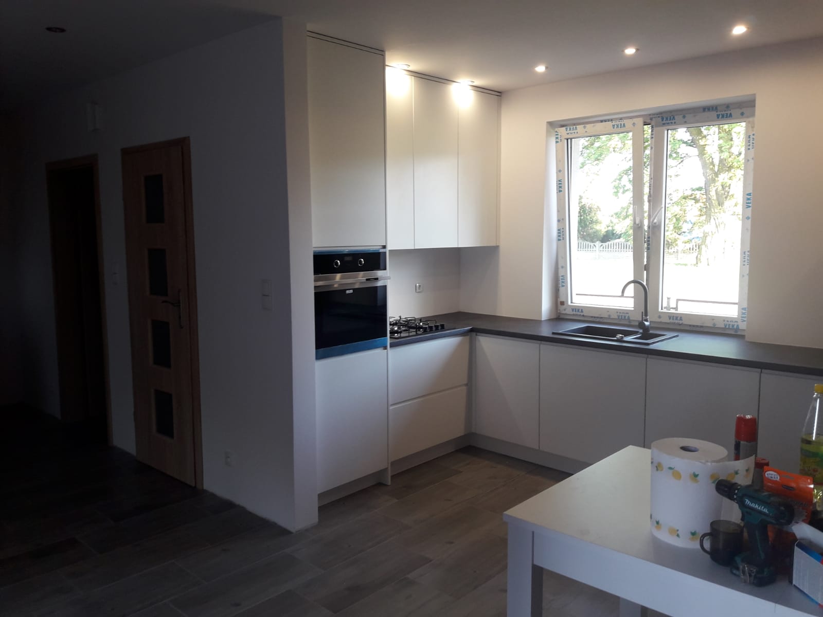 Nowoczesna, jasna kuchnia z białymi frontami szafek, czarnym blatem i oknem z widokiem na zieleń. Na pierwszym planie biały stół z rolką papieru, wiertarką i innymi akcesoriami.