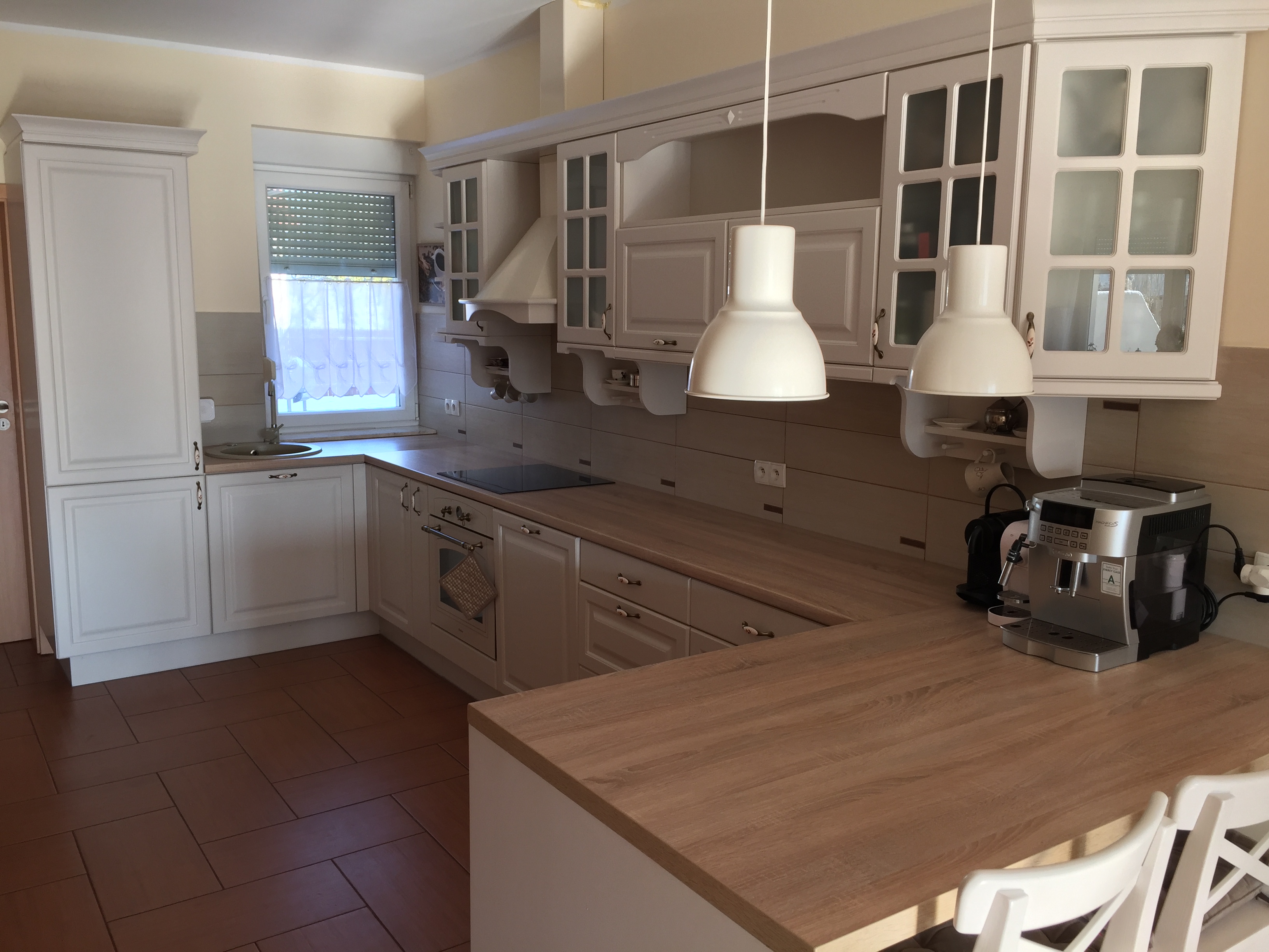 Jasna kuchnia w stylu klasycznym z drewnianym blatem, białymi szafkami i wiszącymi lampami, ekspres do kawy na blacie.