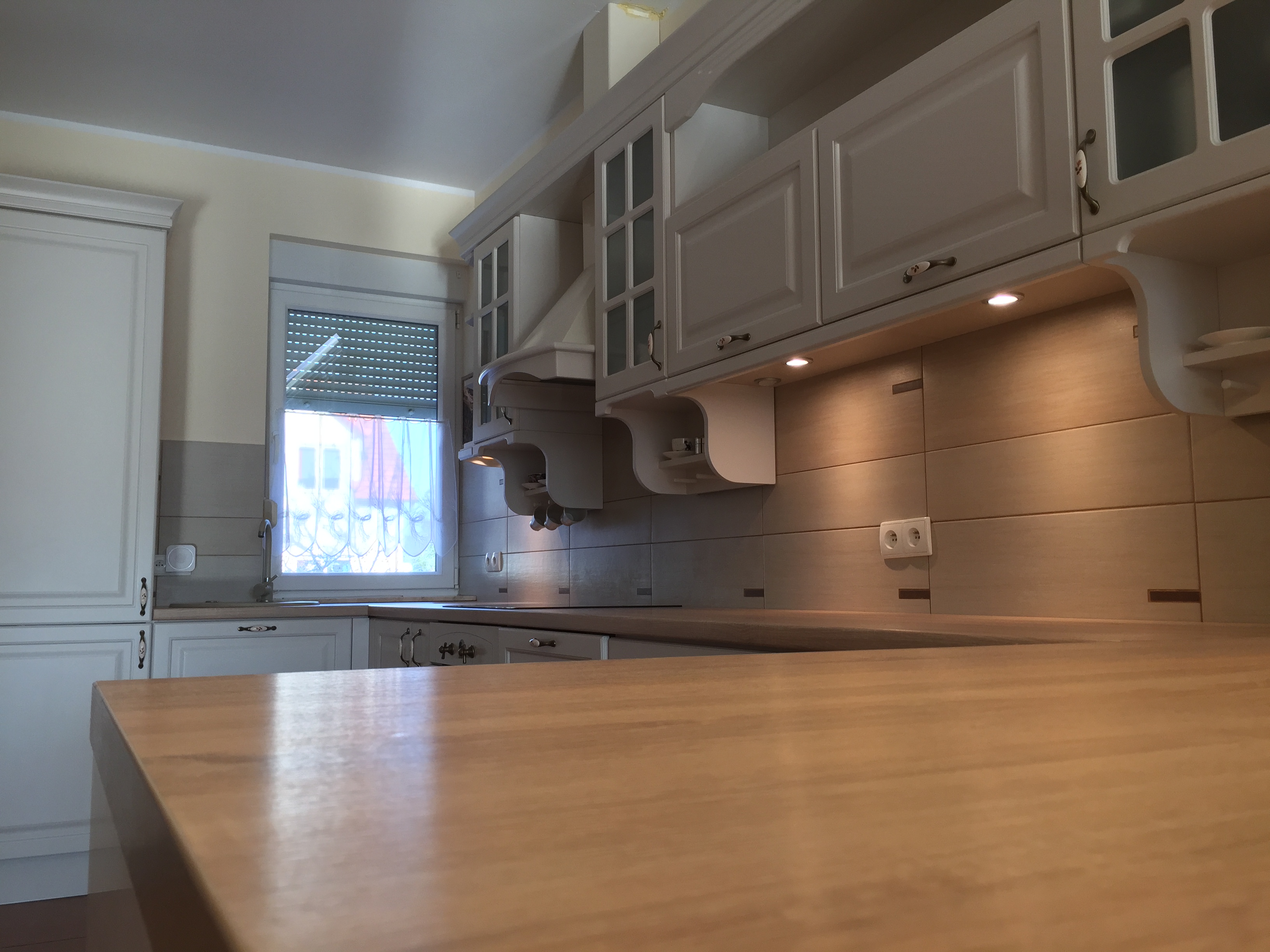 Jasna kuchnia z drewnianym blatem, białymi szafkami z ozdobnymi uchwytami i okapem, widok na okno z roletami.