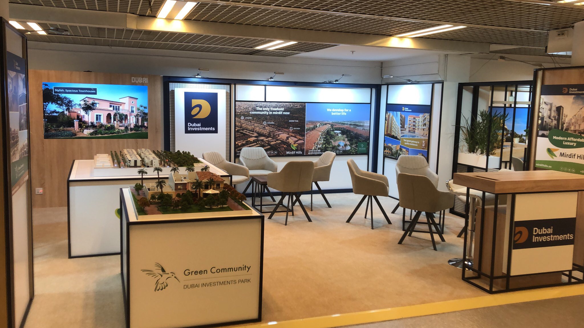 Wnętrze stoiska targowego Dubai Investments z modelami osiedli mieszkaniowych, krzesłami dla klientów i ekranami prezentującymi wizualizacje.