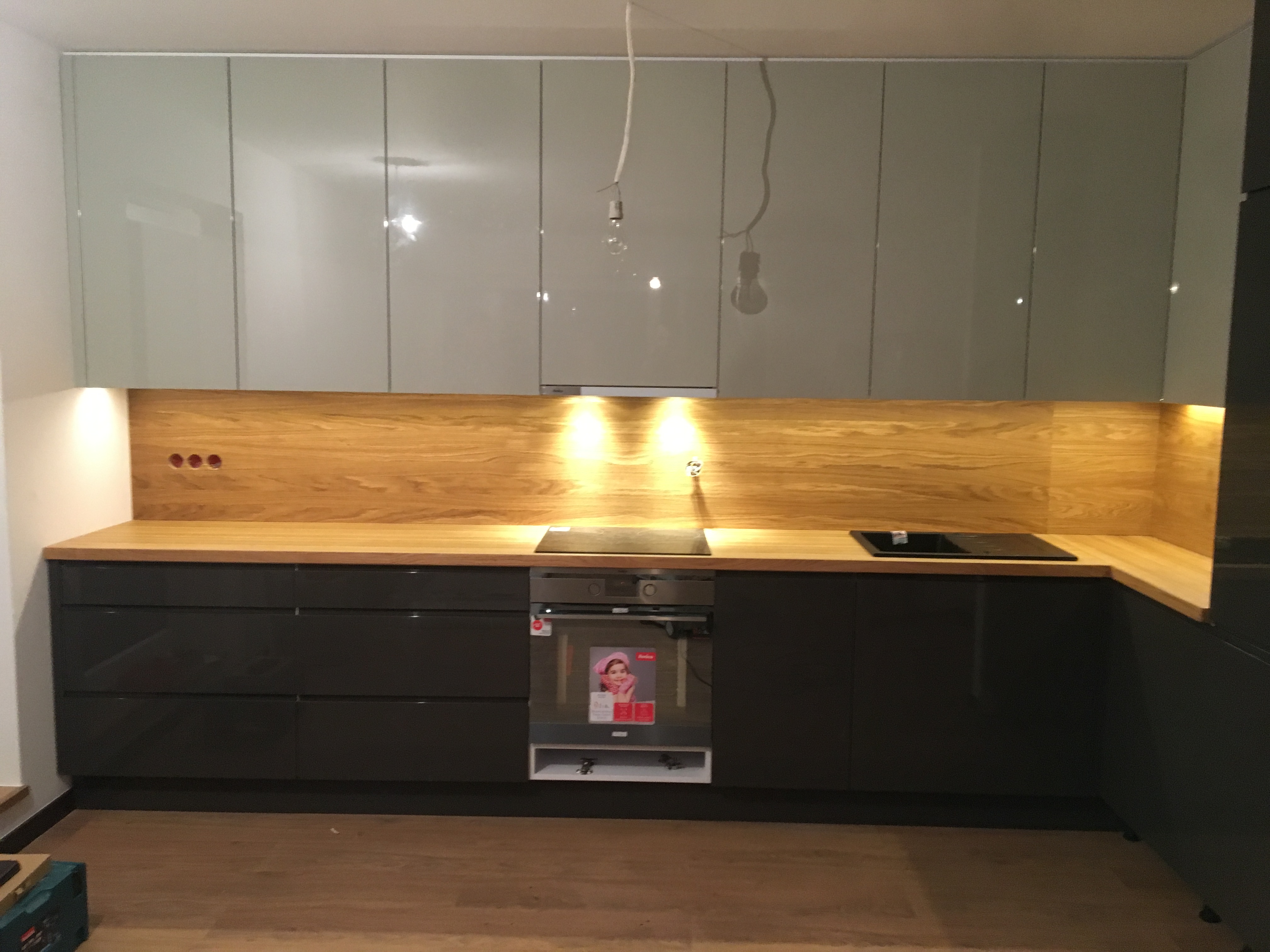 Nowoczesna kuchnia z szarymi, błyszczącymi szafkami górnymi i ciemnymi dolnymi, drewniany blat oraz panel nad blatem. Dwie wiszące żarówki bez kloszy.