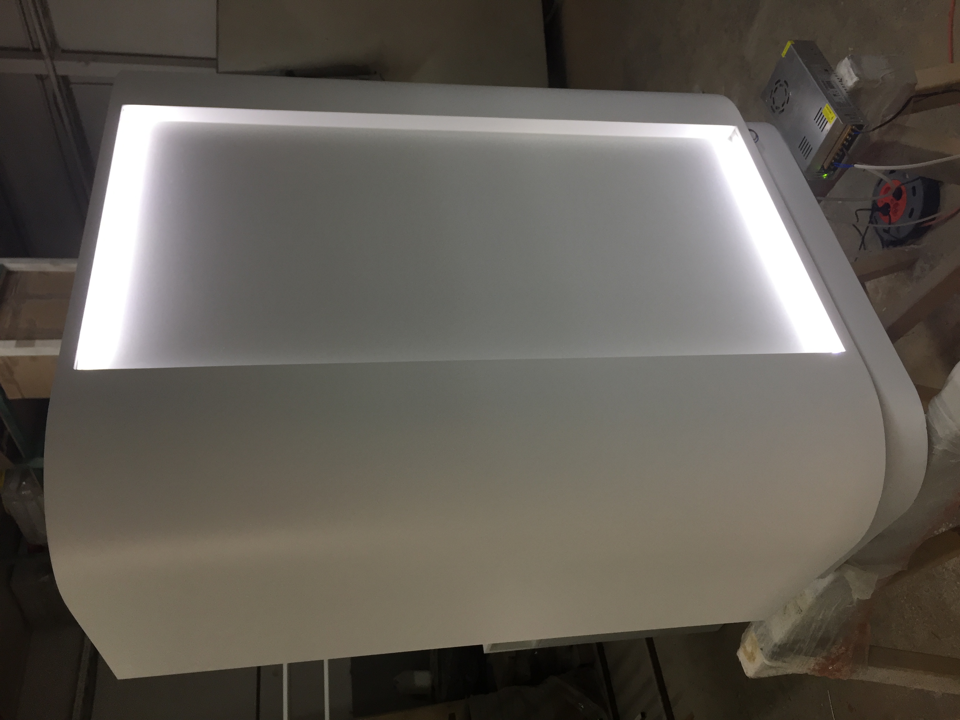 Biała, podświetlana od wewnątrz lada recepcyjna z zaokrąglonymi bokami, widoczny zasilacz i okablowanie w tle.