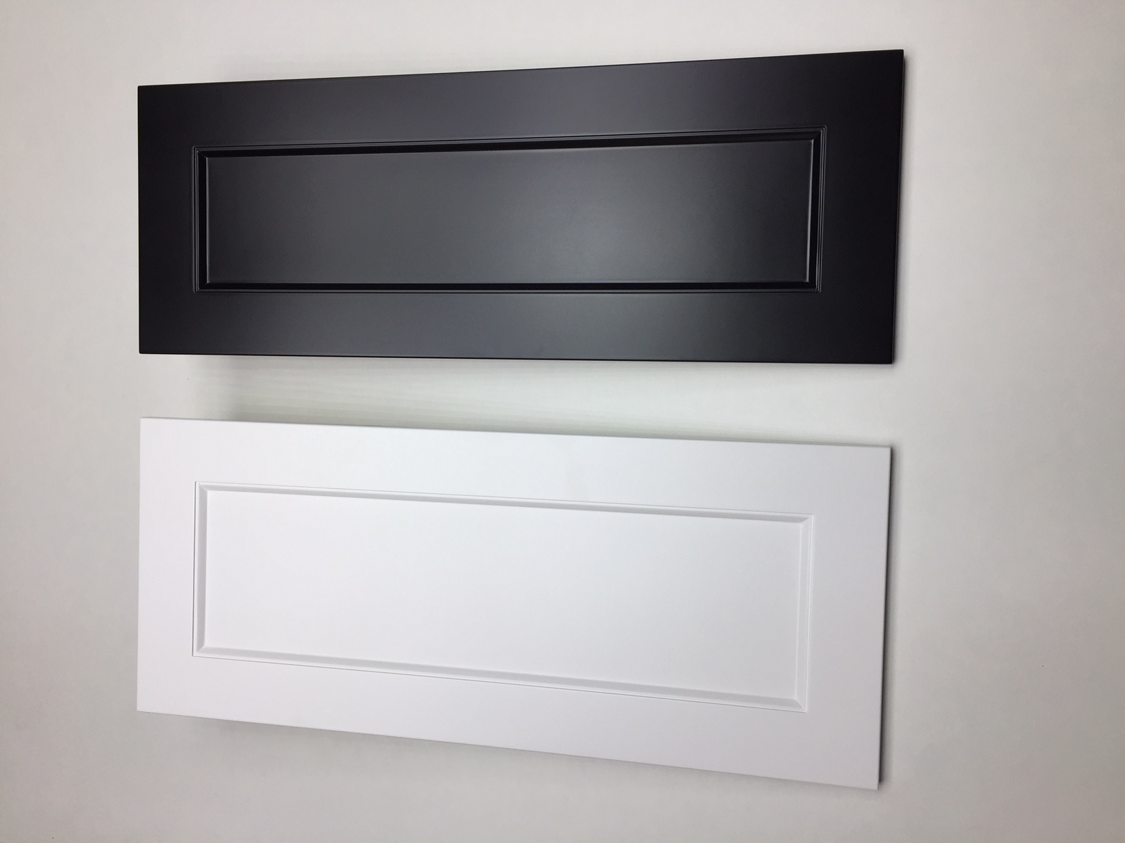 Dwie próbki frontów meblowych: czarny matowy i biały z frezowanym obramowaniem, prezentowane na jasnym tle ściany.