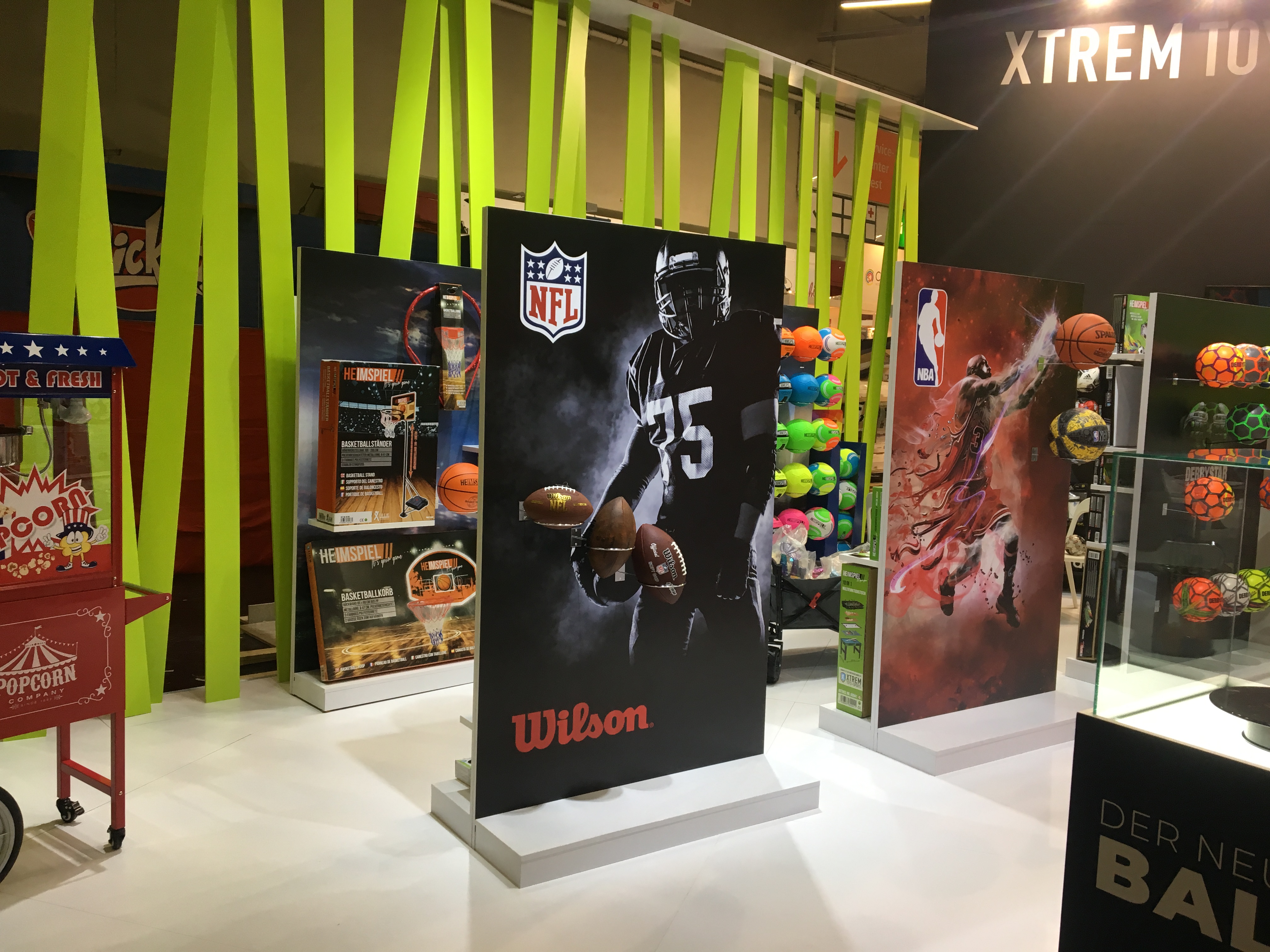 Ekspozycja w sklepie sportowym z planszami reklamowymi przedstawiającymi sportowców i sprzęt sportowy, w tle neonowe zielone dekoracje i automat do popcornu.