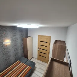 Pokój po remoncie z szarą ścianą o fakturze, oświetleniem punktowym, drewnianymi meblami i sofą z kolorowym pledem.